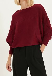 Pull en maille bordeaux avec un col large, une coupe ample et un motif texturé, associé à un pantalon noir taille haute.