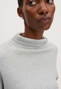 Maglione grigio chiaro con colletto alto a risvolto e texture a costine. Modello che indossa un piccolo orecchino a cerchio d'argento. Inquadratura ravvicinata che mette a fuoco il collo.