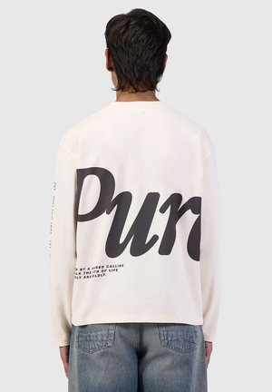 Young person with dark hair wears a white long-sleeve shirt with large black text "Pur" on the back and blue denim jeans, standing facing away.