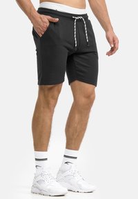 Mann in schwarzen Joggingshorts mit Kordelzug, weißen Sportsocken mit schwarzen Streifen und weißen Sneakers, steht mit einer Hand in der Tasche.