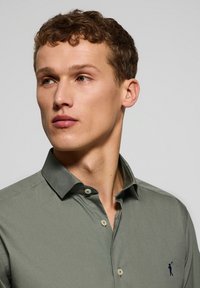 Camicia verde oliva con collo classico, abbottonatura frontale e un piccolo ricamo del logo blu navy sul lato sinistro. Tessuto di alta qualità.