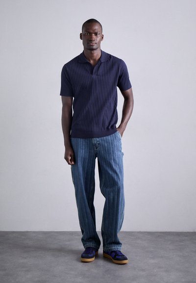 Polo shirt côtelé bleu marine avec un col, associé à un pantalon large à rayures bleu clair. Le modèle porte des baskets violettes et beiges.