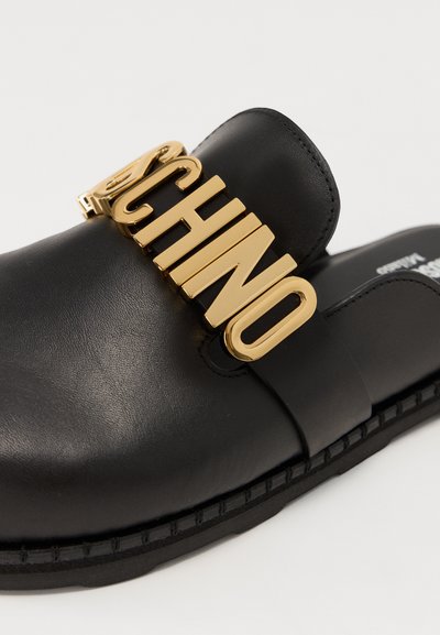 Μαύρη δερμάτινη παντόφλα με φαρδύ λουρί που διαθέτει μεγάλο χρυσό μεταλλικό λογότυπο. Λείος υφής και επίπεδη καουτσούκ σόλα για εύκολη χρήση.