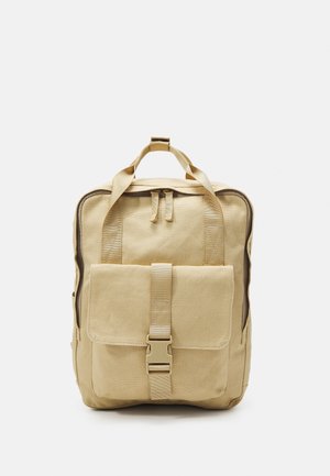 Sac à dos en toile beige avec poche avant à boucle, poignée supérieure et fermeture éclair, présenté sur fond blanc.