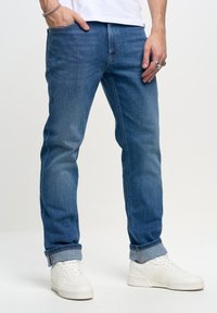 Jeans in denim blu con una vestibilità classica, leggermente sfumati e con orli arrotolati. Indossati con sneakers bianche, mostrano uno stile informale.