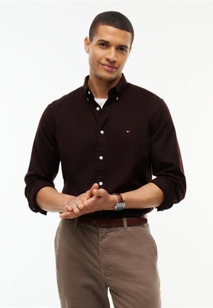 Homme portant une chemise boutonnée marron foncé et un pantalon beige, les mains jointes devant, avec un léger sourire sur fond blanc.