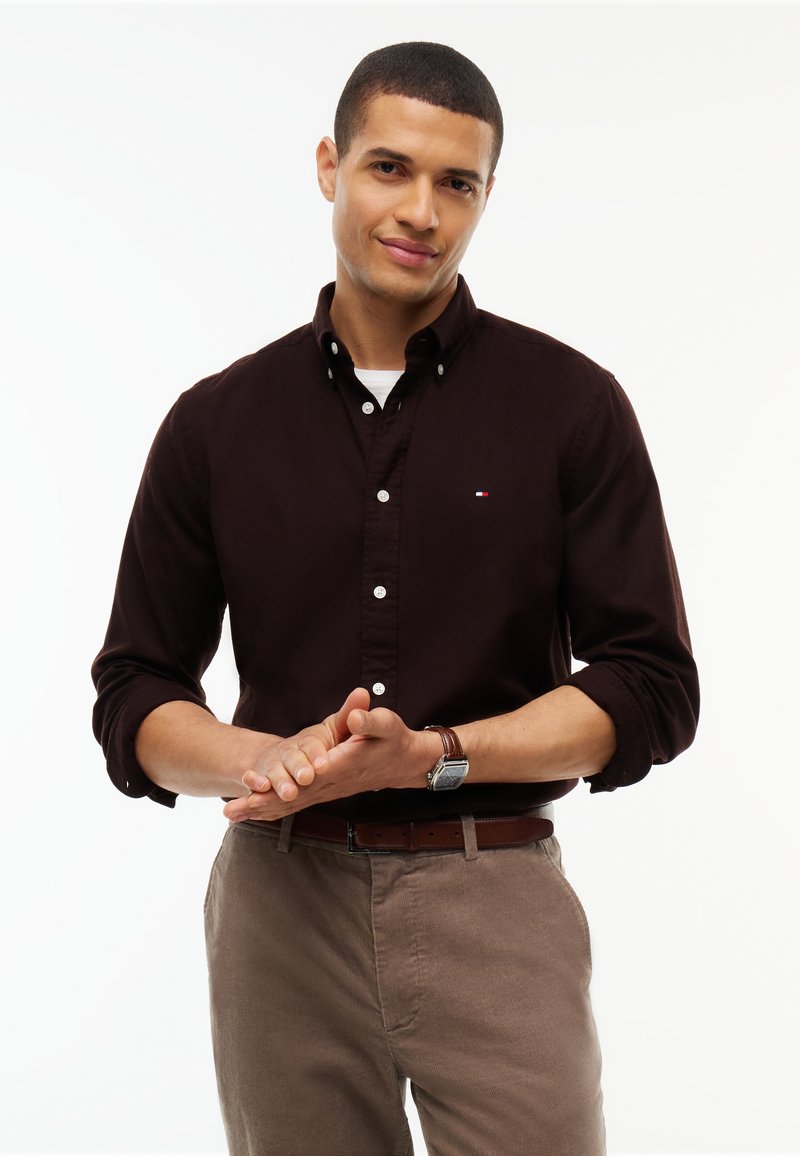 Homme portant une chemise boutonnée marron foncé et un pantalon beige, les mains jointes devant, avec un léger sourire sur fond blanc.