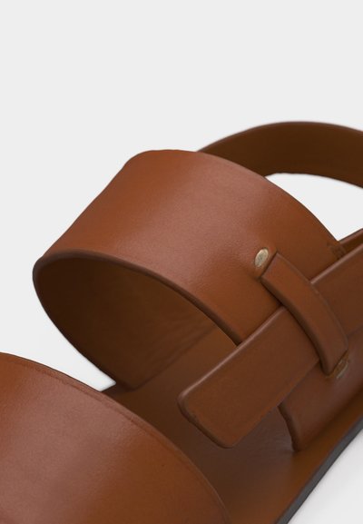 Κοντινό πλάνο καφέ δερμάτινου σανδαλιού με φαρδιά μπροστινή λουρίδα και ρυθμιζόμενο πλαϊνό λουρί με μεταλλικό καρφί, σε λευκό φόντο.