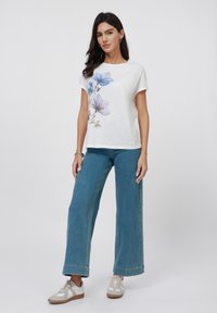 T-shirt blanche avec imprimé floral bleu, à manches courtes, portée avec un jean bleu à jambes larges et des baskets grises. Design simple et décontracté avec un tissu léger.