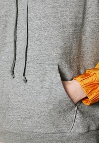 Haut à capuche gris avec cordons, doté d'une poche frontale. Manchons en tissu orange brillant avec un design froncé, affichant des textures contrastées.