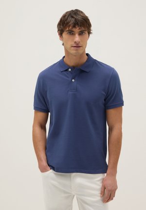 Hombre con camiseta tipo polo azul de manga corta y pantalones blancos de pie frente a un fondo claro y liso con una mano en el bolsillo.