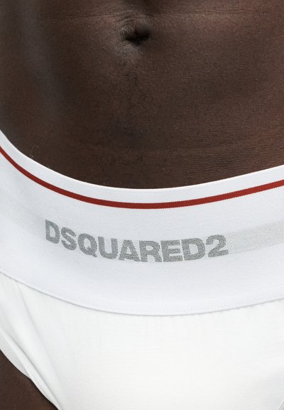 Κοντινό πλάνο στην κοιλιά ενός ατόμου που φοράει λευκά εσώρουχα με κόκκινη λωρίδα και λογότυπο "DSQUARED2" στη μέση.