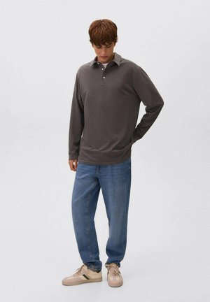 Giovane uomo che guarda in basso, indossando un polo a maniche lunghe grigio scuro, jeans blu e sneaker beige con strisce verdi, in piedi su uno sfondo bianco.