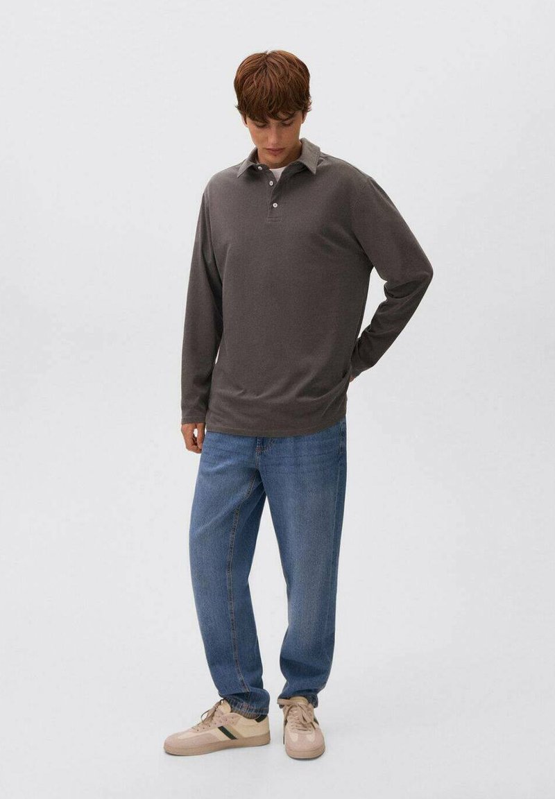 Junger Mann, der nach unten schaut, trägt ein dunkelgraues Langarmpolo, eine blaue Jeans und beiges Sneakers mit grünen Streifen, stehend vor einem weißen Hintergrund.