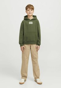Sudadera verde con un bolsillo frontal y etiqueta de logo, combinada con pantalones cargo beige y zapatillas. La sudadera tiene un corte relajado y capucha ajustable.