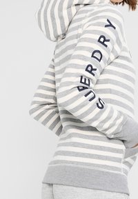 Persona che indossa un hoodie a righe grigie e bianche con il testo "SUPERDRY" sulla manica, mani nelle tasche, mostrata di lato.
