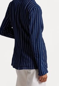 Person wearing a navy blue pinstripe jacket and white pants, shown from the back with one hand resting near their hip.