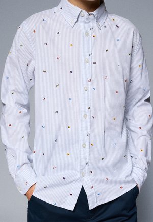 Biała koszula zapinana na guziki z cienkimi jasnoniebieskimi paskami i małymi, wielokolorowymi haftowanymi flagami morskimi rozmieszczonymi na całej powierzchni, noszona z ciemnymi spodniami.