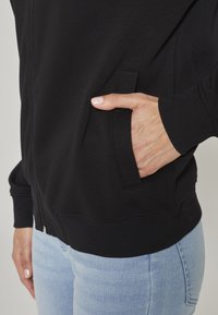 Sweat à capuche zippé noir avec poignets et ourlet côtelés. Comprend une poche latérale, une texture lisse, et une coupe ajustée, porté avec un jean bleu clair.