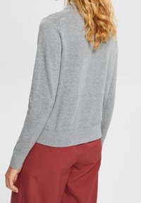 Maglione grigio con colletto alto, polsini e fondo a coste, e una texture morbida. Abbinato a pantaloni borgogna, che mostrano un design di abbigliamento casual.