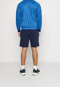 Blå nylon bomberjacka med ribbade ärmslut, kombinerad med marinblå shorts. Personen bär svarta sneakers med gröna accenter och vita strumpor.