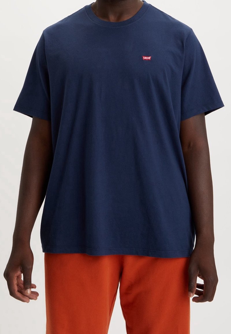 Personne à la peau foncée portant un t-shirt Levi's bleu marine uni et un pantalon orange vif, debout avec les bras détendus le long du corps.