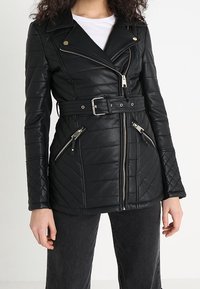 Chaqueta de motociclista de cuero negro acolchada con cremalleras plateadas, cintura con cinturón y solapas grandes. Cuenta con dos bolsillos con cremallera y un ajuste estructurado.