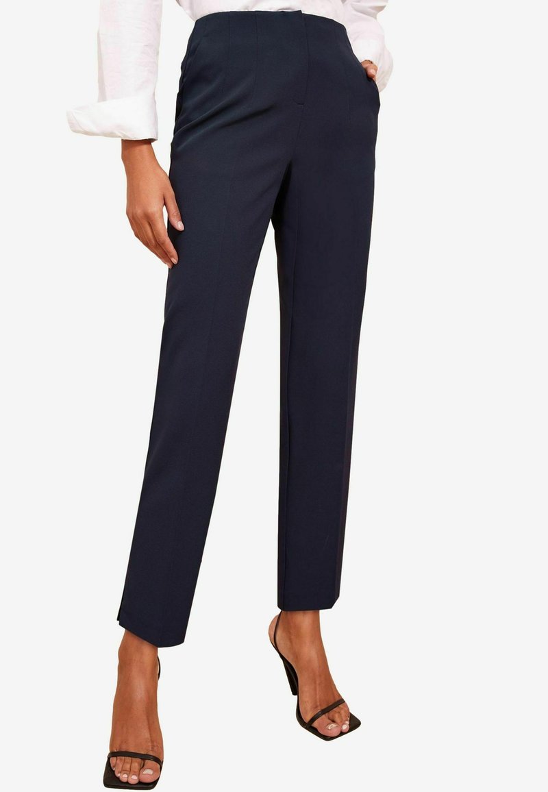 Mørkeblå skræddersyede bukser med lige ben, sidelommer og lette plisseringer. Stoffet ser glat og struktureret ud. Bæres med sorte sandaler.