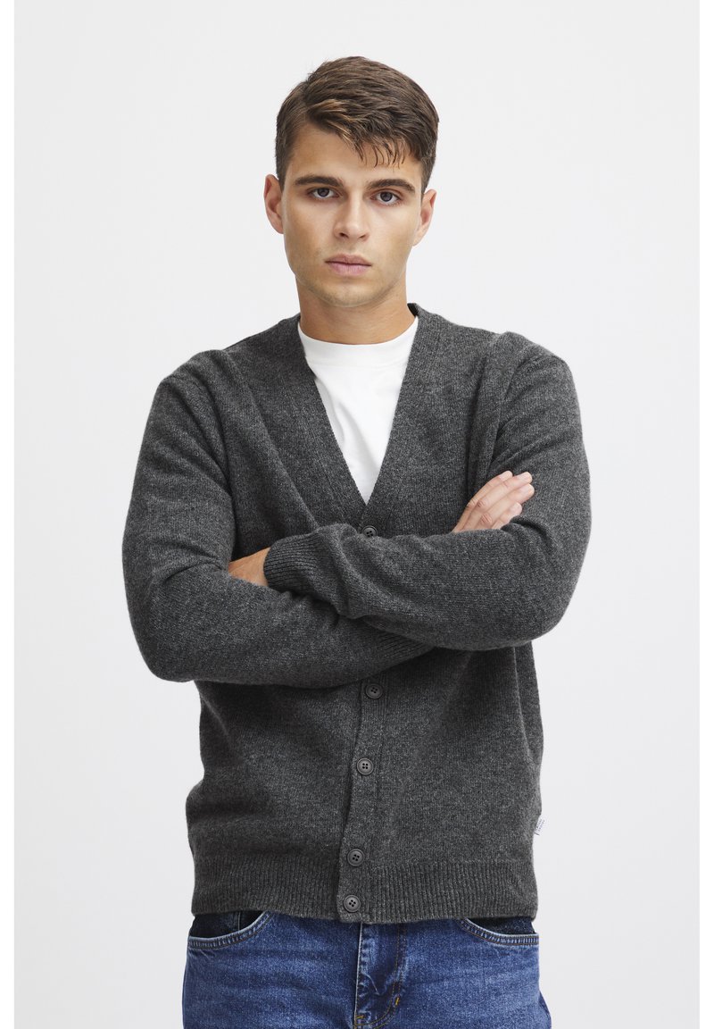 Cardigan de punto gris oscuro con escote en V, cierre de botones, puños y dobladillo acanalados; llevado sobre una camisa blanca. Textura ligera, corte casual.