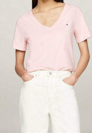 Personne portant un t-shirt rose clair à col en V et manches courtes, rentré dans un pantalon blanc taille haute, les mains dans les poches.