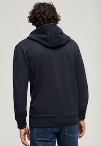 Mørkeblå hættetrøje lavet af bomuld med en tekstureret overflade, med en hætte med snore og ribbede manchetter, båret over denimjeans.