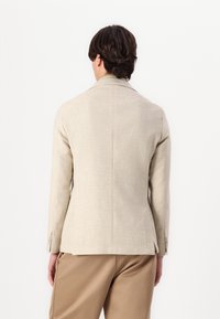 Veste ajustée beige clair avec une finition texturée, design à un bouton, et fentes latérales, associée à un pantalon marron clair.