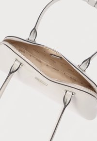 Λευκό δερμάτινο τσάντα χειρός με δύο λαβές, ασημένιο φερμουάρ και μπεζ εσωτερική επένδυση με τυπωμένα λογότυπα "GUESS".