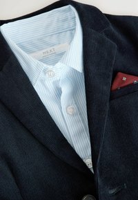 Námornícky kordový blejzer s bledomodrou pruhovanou košeľou s gombíkmi pod ním. Obsahuje červený vreckový šál z patterned látky.
