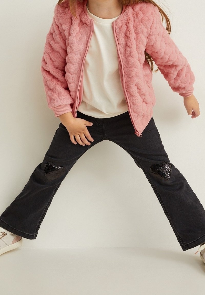 Roze donzen bomberjack met ribbelmanchetten, witte T-shirt, zwarte spijkerbroek met zwarte pailletten hartapplicaties op de knieën, en witte schoenen.