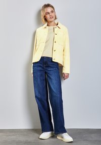 Ljusgul rutig jacka med stora knappar, lagd över en beige tröja och buren med blå vida jeans och vita sneakers.