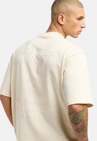 Cream-colored oversized t-shirt featuring embossed graphics, including "CARLO COLUCCI" and a stylized logo, with a soft cotton texture.