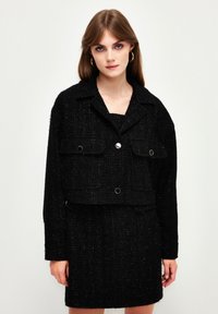 Schwarze Tweed-Cropped-Jacke mit zwei vorderen Taschen, goldenen Knopfakzenten und einer leicht strukturierten Oberfläche, kombiniert mit einem passenden Rock.