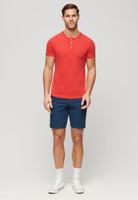 T-shirt henley rouge à manches courtes avec patte de boutonnage à trois boutons, associé à un short bleu. Mod�èle portant des baskets blanches et des chaussettes blanches texturées.