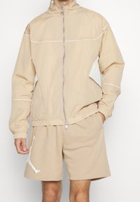 Beige lättviktig jacka med vita accenter och dragkedjefickor, med hög krage och elastiska muddar, tillsammans med matchande shorts.