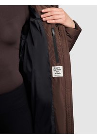 Brązowa pikowana kurtka z czarną podszewką, pokazana rozpięta. Posiada zamek błyskawiczny, poziome przeszycia oraz metkę z napisem "Ciepły uścisk wykonany z tkaniny".