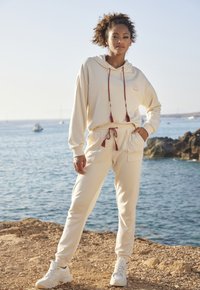 Set di felpa e pantaloni della tuta color crema con lacci regolabili, tasche e una grafica del sole. Indossato con scarpe da ginnastica bianche vicino al mare.