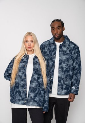 A man and woman stand side by side wearing matching blue camouflage jackets, white shirts, and black pants against a plain background.