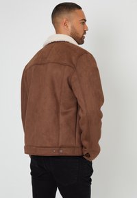 Brun faux suede jakke med en hvid shearling krave, der har en lige pasform, sidesøm og diskrete metaldetaljer ved kanten.