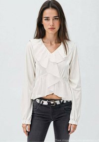 Blusa bianca con maniche lunghe e rouches sul colletto e davanti, in tessuto strutturato, abbinata a jeans neri e una cintura fantasia.
