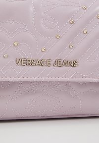 Lawendowa skórzana torba na ramię z wytłaczanym wzorem i złotym okuciem z logo "VERSACE JEANS" z przodu. Gładka tekstura.