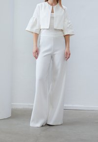 Chaqueta blanca recortada con mangas anchas, llevada sobre una blusa blanca y pantalones de pierna ancha de cintura alta. Textura suave, diseño minimalista.