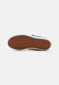 Suela de una zapatilla con un fondo de goma marrón con patrón, entresuela blanca y toques de azul en la parte superior, con un acento rojo.