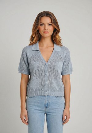 Donna che indossa un cardigan a maniche corte color azzurro pallido con motivi floreali e bottoni, abbinato a jeans azzurri, su uno sfondo semplice.