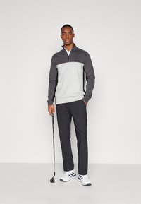 Grå och vit halvdraget pullover med logotyp, matchat med mörka byxor och vita golfskor. Modellen håller en golfklubba och står mot en enfärgad bakgrund.